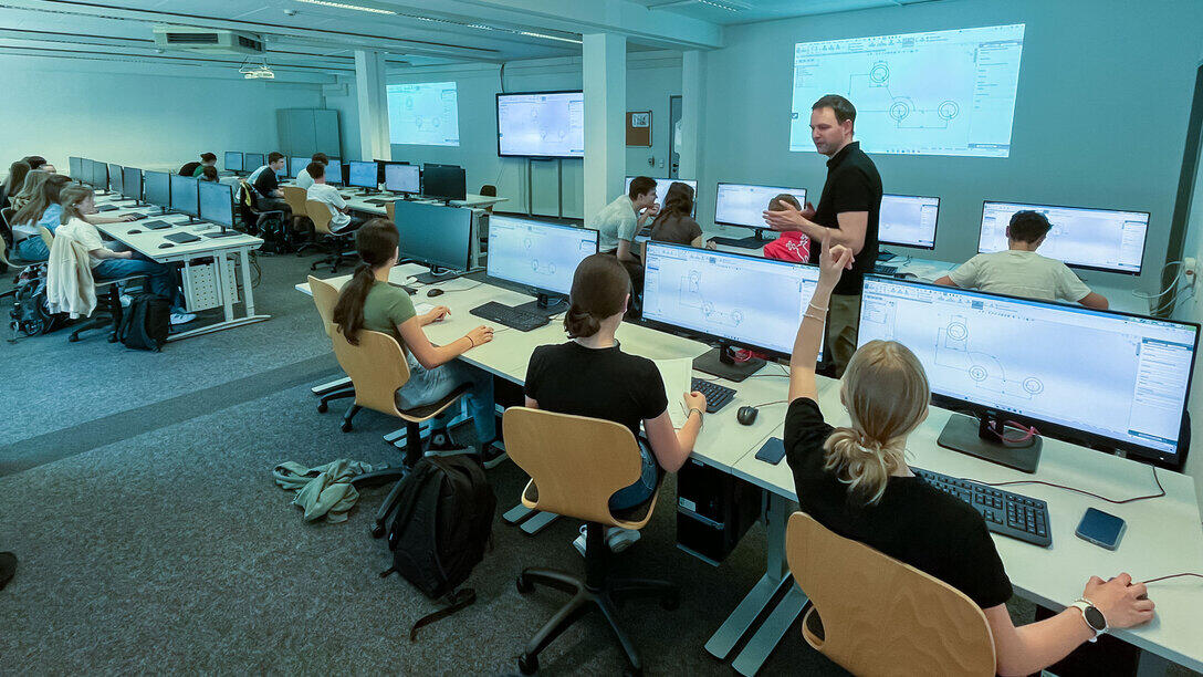 Blicke in einem Computerpool. Vor den Rechnern mit großen breiten Monitoren sitzen Schüler:innen. Ein männlicher Dozent geht erklärend durch die Reihen.