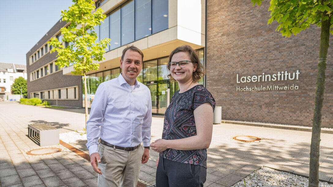 Eine männliche udn eine weibliche Person stehen nebeneinander und gestikulieren freundlich. Im Hinterggrund ist iein verklinkertes Gebäude mit dem Schriftzug "Laserinstitut Hochschule Mittweida" zu sehen.