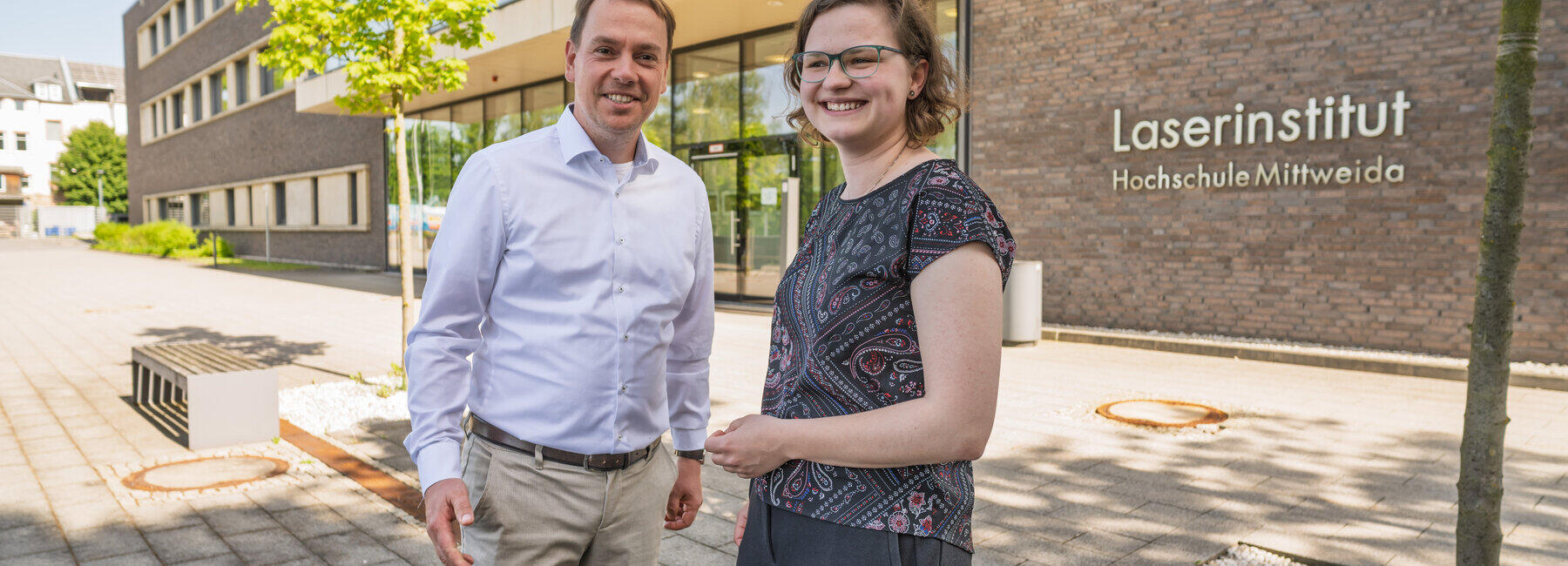 Eine männliche udn eine weibliche Person stehen nebeneinander und gestikulieren freundlich. Im Hinterggrund ist iein verklinkertes Gebäude mit dem Schriftzug "Laserinstitut Hochschule Mittweida" zu sehen.