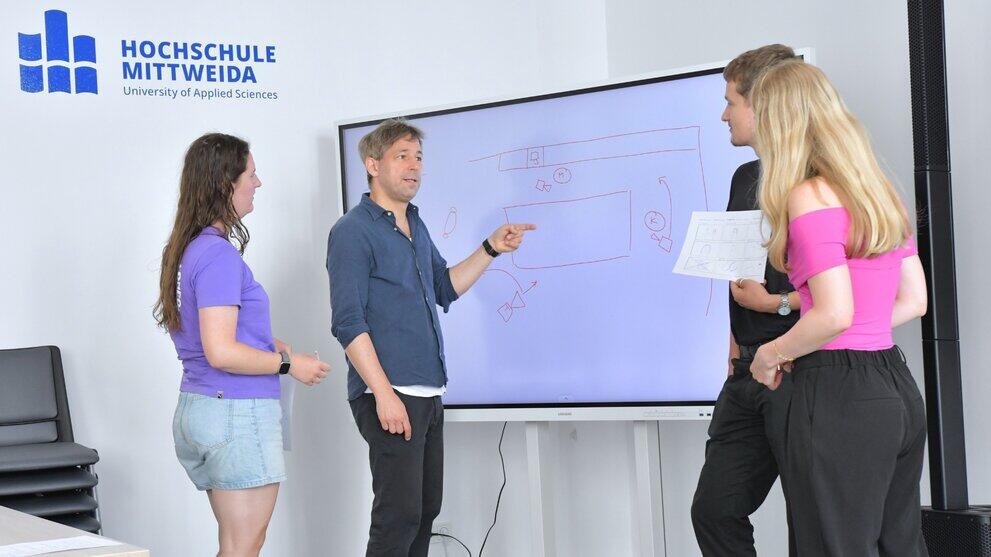 Several students and a lecturer are standing in front of a digital whiteboard, discussing a sketch.