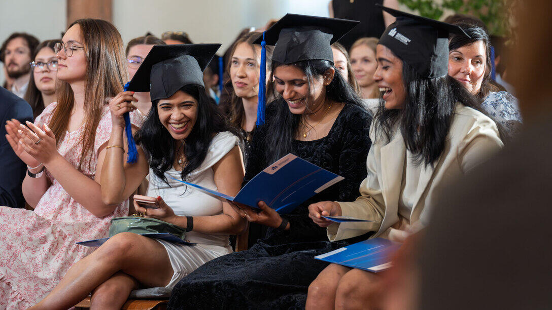 Drei weibliche Graduierte sitzen mit Absolvent:innenhut nebeneinander und lachen