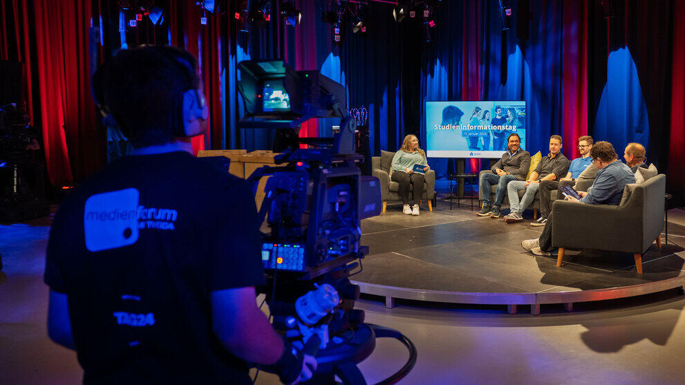 A view into the TV studio, past a studio camera, of people sitting on sofas on a low platform.