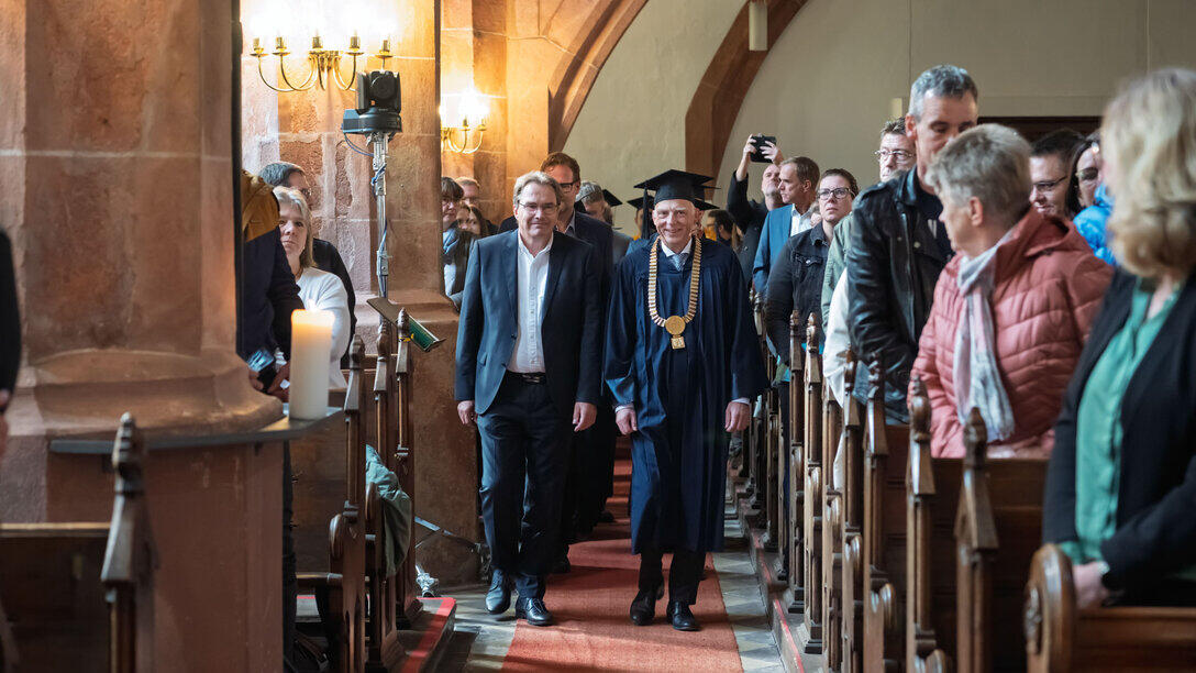 Professor:innen und andere Personen beim Einzug durch den Gang im Kirchenschiff. Die Besucher:innen stehen.