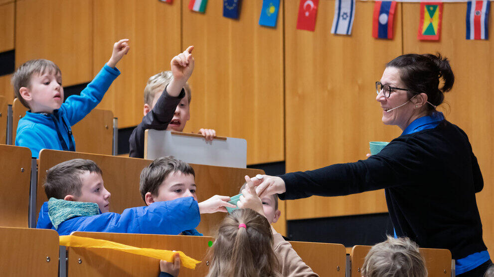 Dozentin reicht Kindern in einem Hörsaal mit Holzwänden und internationalen Flaggen etwas, mehrere Kinder heben die Hand.