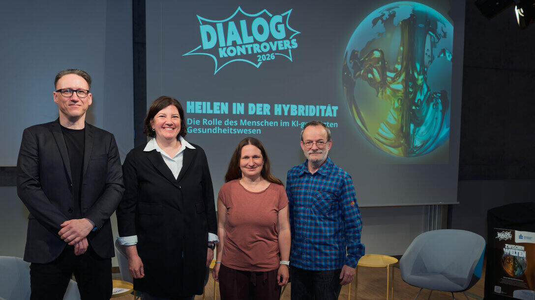 Zwei weibliche und zwei männliche Personen stehen lächelnd vor der Leinwand hinter ihnen, die das Logo des Dialog Kontrovers und das aktuelle Thema „Heilen in der Hybridität. Die Rolle des Menschen im Kl-gestützten Gesundheitswesen“ zeigt.