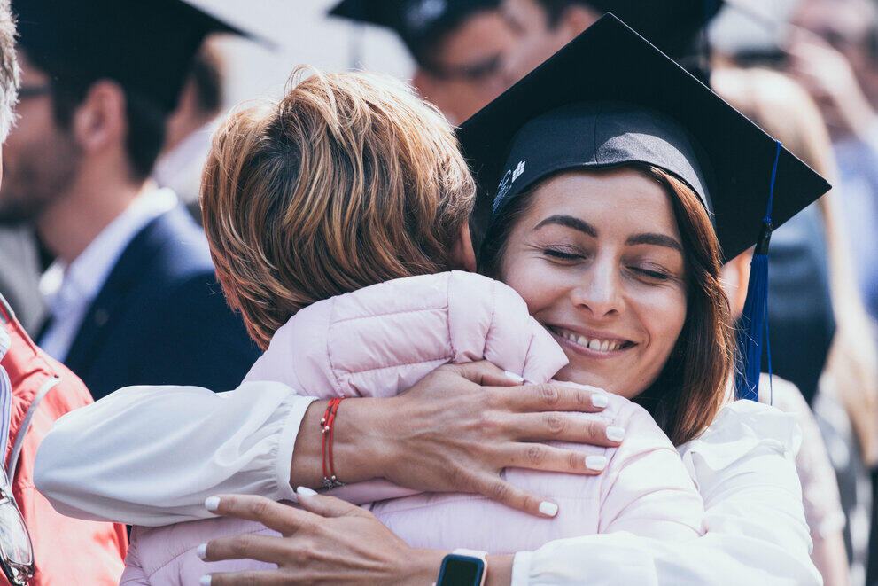 Alumna mit Hut umarmt Familienmitglied mit Lachen im Gesicht
