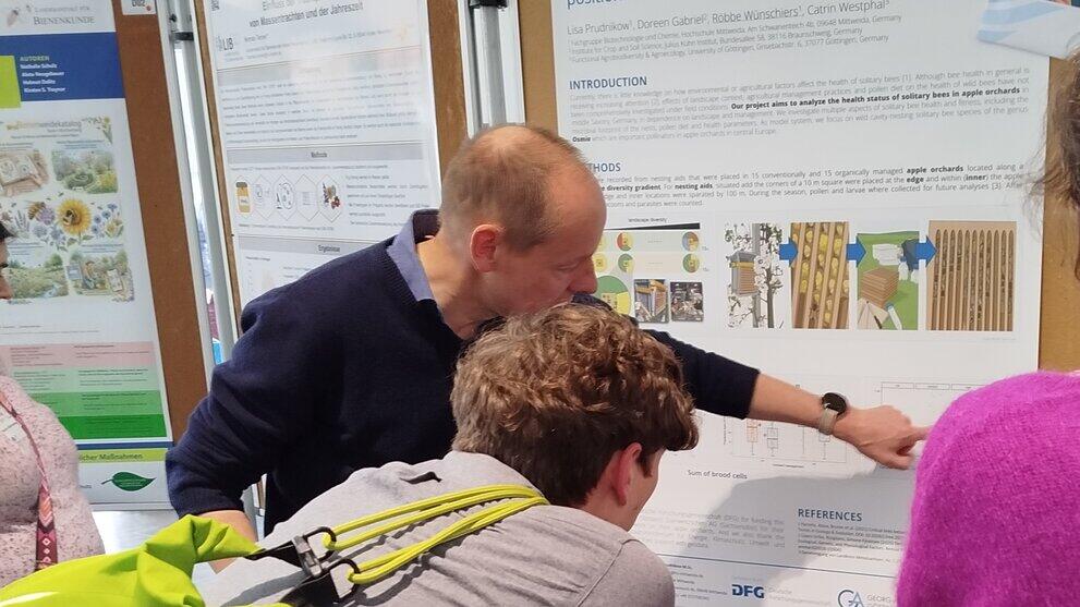 Drei Personen stehen vor einer Posterwand, eine Person zeigt erklärend auf ein Bild