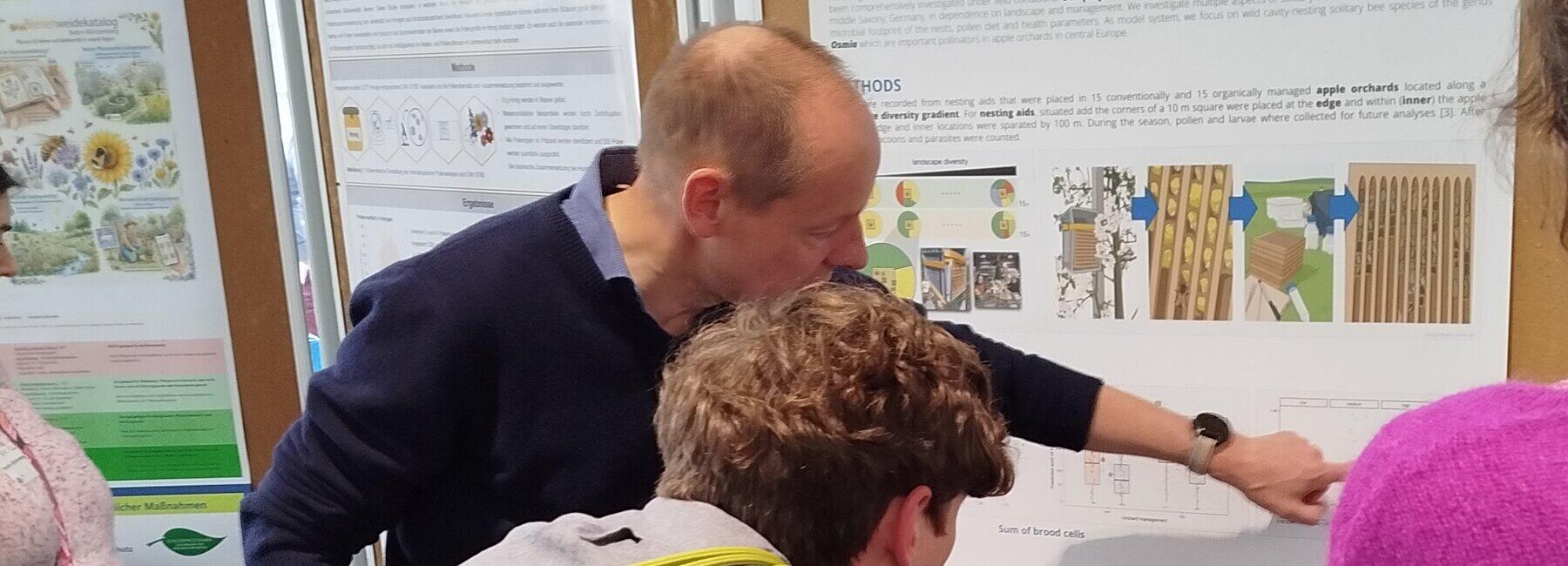 Drei Personen stehen vor einer Posterwand, eine Person zeigt erklärend auf ein Bild