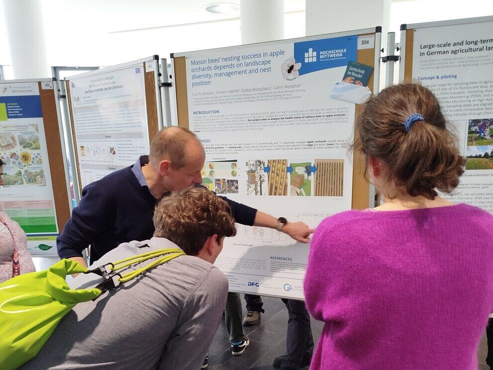 eine Person erklärt ertwas anhand eines Poster zwei Personen hören zu