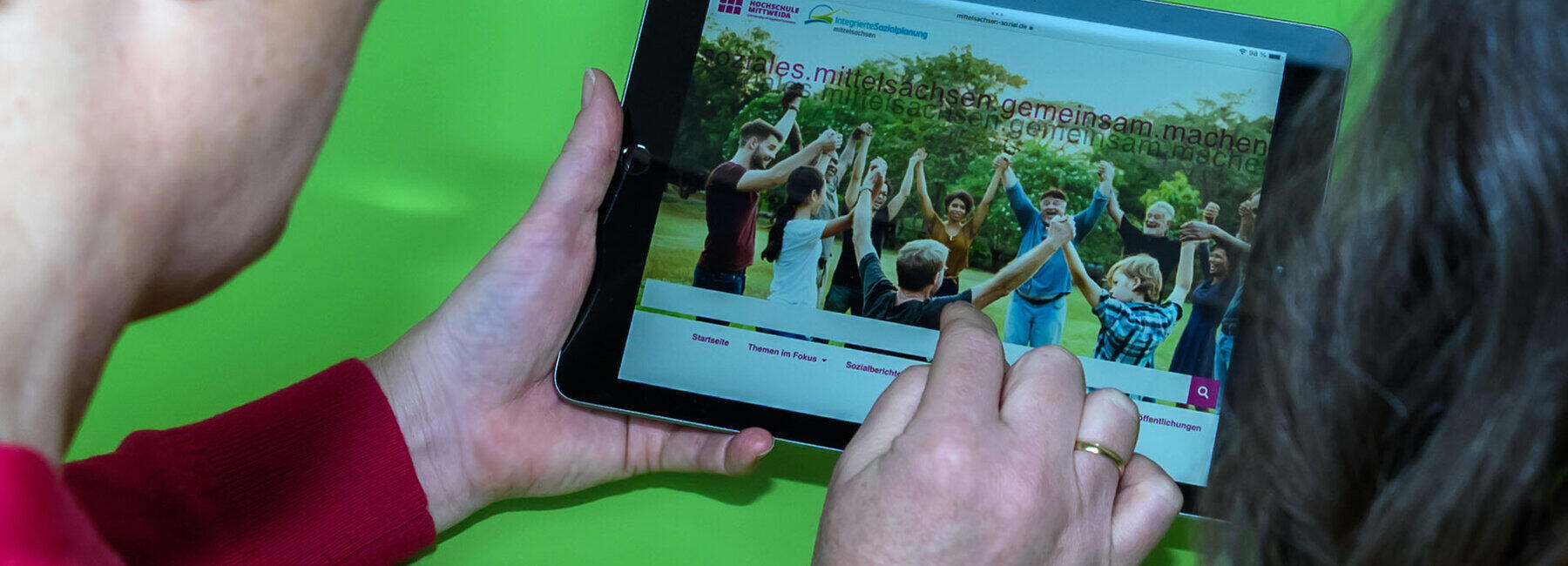 Blick über die Schultern zweier Personen auf ein Tablet in den Händen der linken Person. Sie zeigt mit einem Finger der rechten Hand auf die Oberfläche des Tablets. Es zeigt eine Webseite mit einem Bildmotiv mit Menschen unterschiedlichen Geschlechts und Alters, die im Kreis stehend sich an den Händen fassen. Über das Bildmotiv ist zweimal der Schriftzug mit unterschiedlicher Deckkraft „soziales.mittelsachsen.gemeinsam.machen“ gelegt.