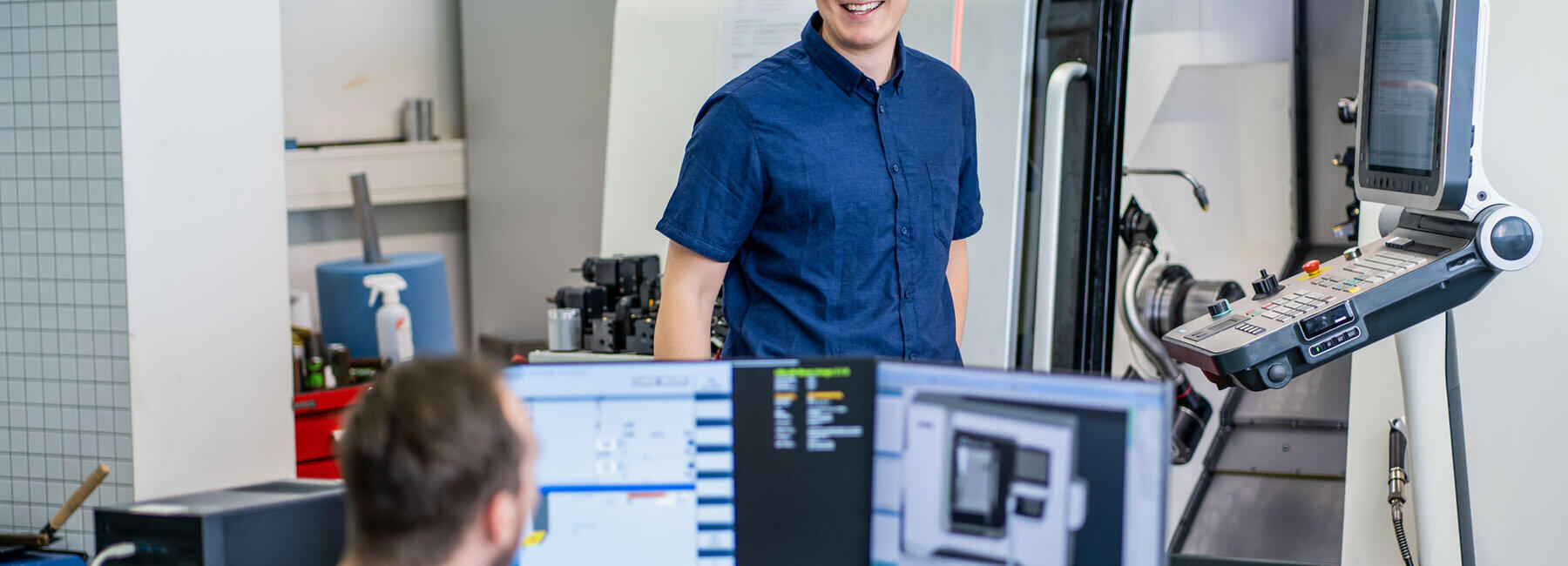 A young man is standing beaming in front of some equipment in the background. In the foreground, another man can be seen sitting in front of some monitors.