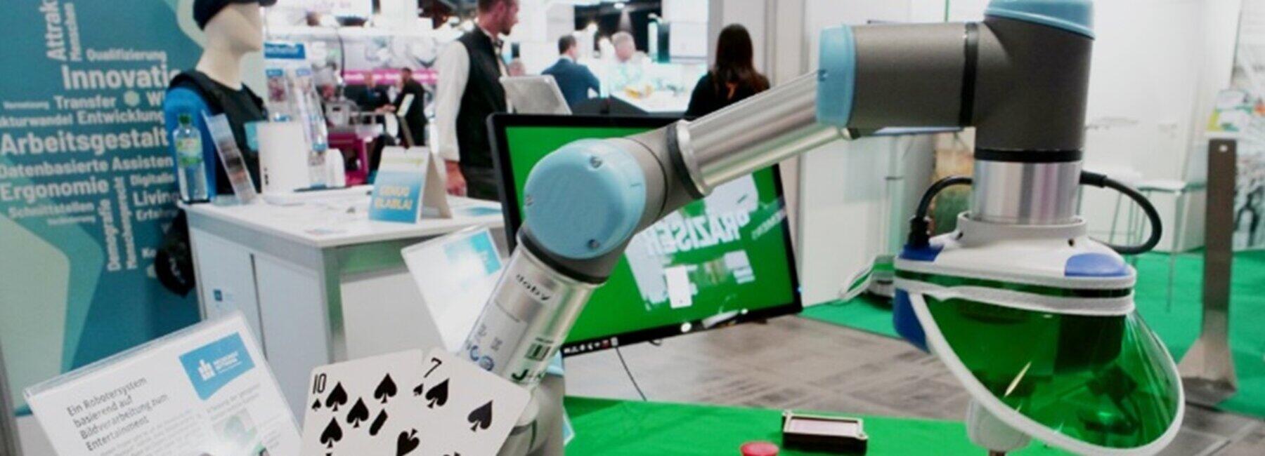 menschliche Hände halten Spielkarten vor einem Roboterarm, im Hintergrund ein Messestand