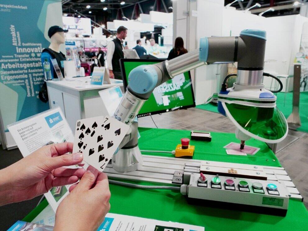 menschliche Hände halten Spielkarten vor einem Roboterarm, im Hintergrund ein Messestand