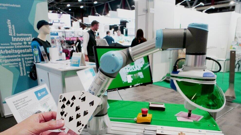 menschliche Hände halten Spielkarten vor einem Roboterarm, im Hintergrund ein Messestand
