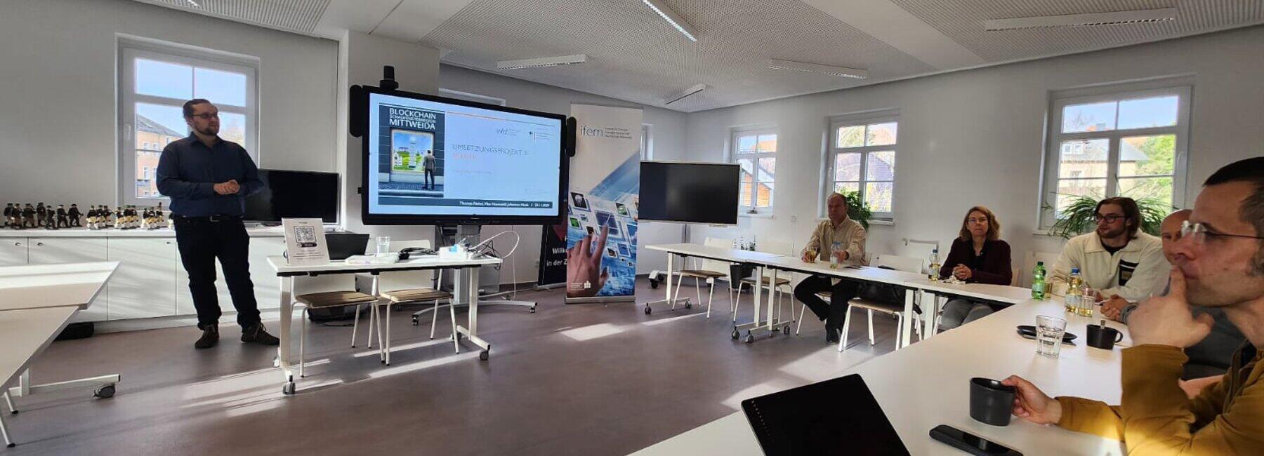 A man is standing in front of a presentation screen; other people are sitting in the room