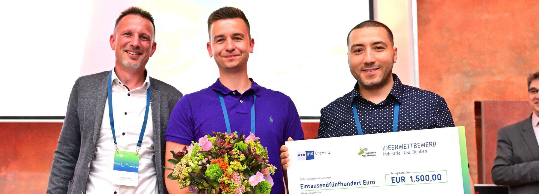 Drei männliche Personen stehen nebeneinander, die mittlere Person hält einen Blumenstrauß, die person ganz rechts eine Urkunde in den Händen