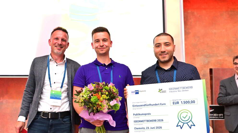 Drei männliche Personen stehen nebeneinander, die mittlere Person hält einen Blumenstrauß, die person ganz rechts eine Urkunde in den Händen