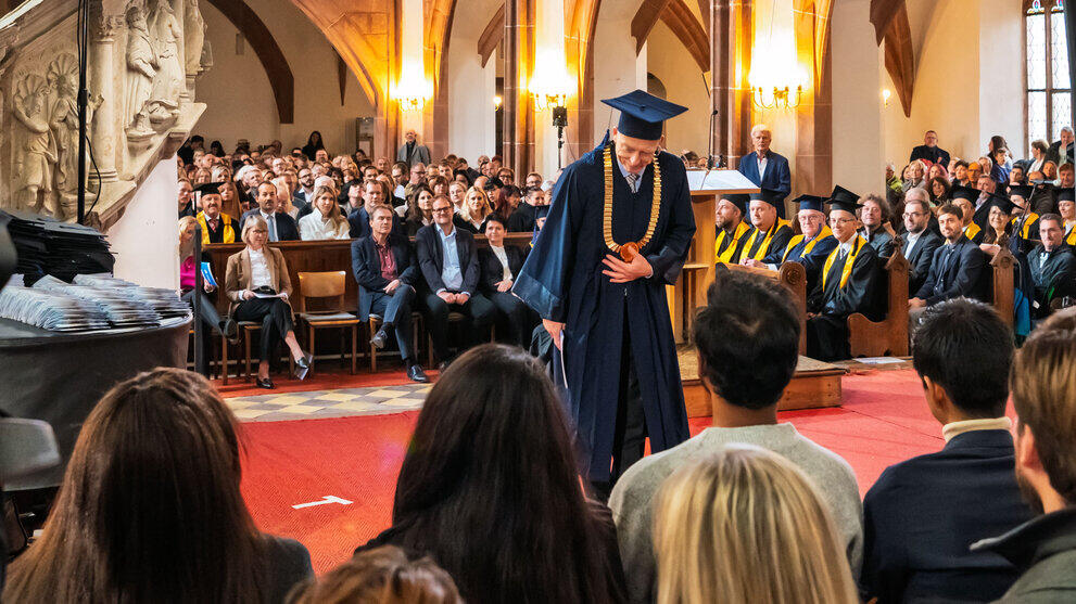 A man in a blue academic gown, wearing a gold chain of office and a doctoral cap, bows to the people seated in the foreground of the picture.