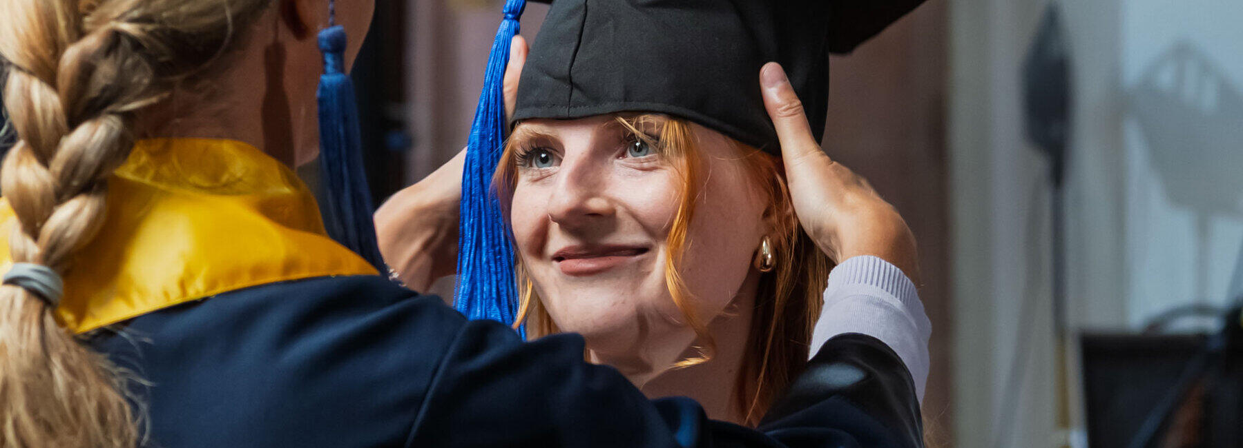 A woman in an academic gown and wearing a doctoral cap places the graduate’s cap on the graduate’s head.