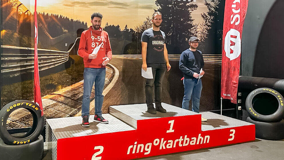Drei junge männliche Personen stehen auf einem dreistufigen Siegerpodest und halten Urkunden in den Händen. Auf dem Podest ist weiß auf rot „Ring Kartbahn“ zu lesen. Rennreifen sind links und rechts vom Podest drapiert. Der Hintergrund ist die Abbildung eines Ausschnitts einer Rennkurve mit Leitplanken.