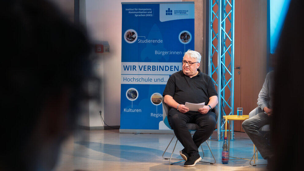 Männliche Person im Sessel auf dem Podium. In Hintergrund steht ein Rollup des IKKS unter anderem mit dem Text "Wir verbinden".