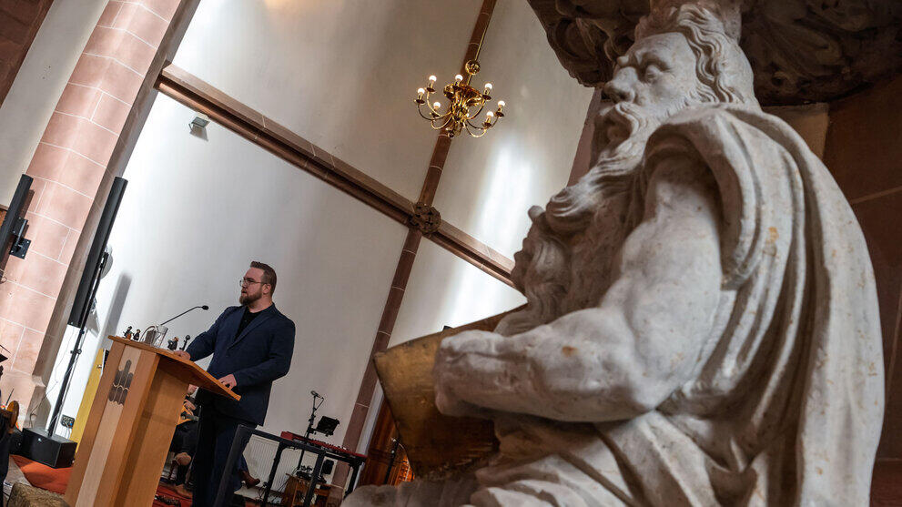 A view of the speaker in the background, seen past a sandstone figure depicting Moses in the foreground. The speaker and the figure share the characteristic of having a full beard