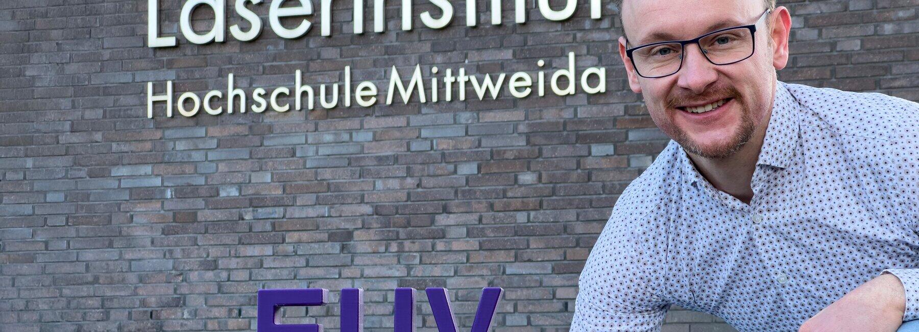 A man is crouching in front of a building wall bearing the inscription ‘Laser Institute, Mittweida University of Applied Sciences’; next to him is a sign reading ‘EUV’