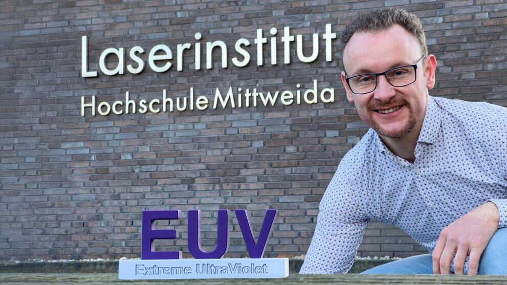 Männliche Person hockt vor Hauswand mit der Aufschrift Laserinstistut Hochschule Mittweida, neben ihm steht ein Schriftzug EUV ein