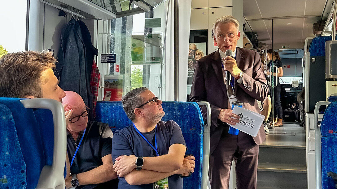 A man holding a microphone is standing on a train and speaking.