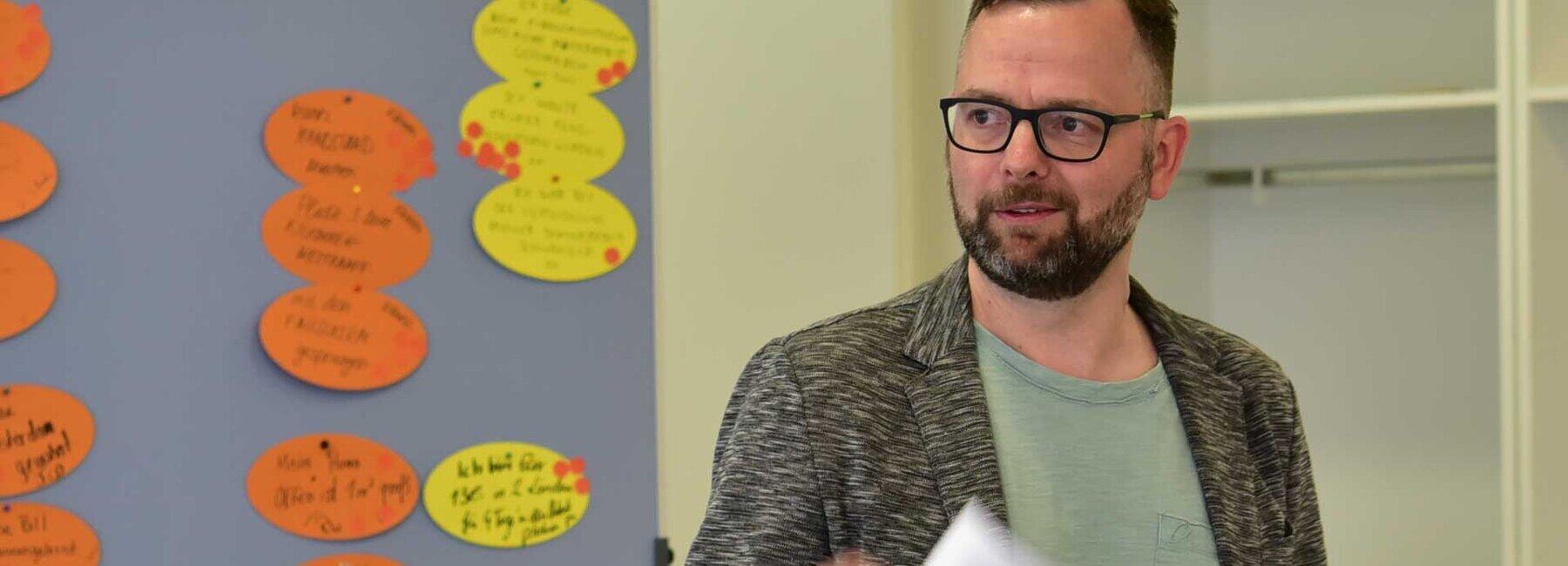 Eine männliche Person steht referierend mit einem Blatt Papier in der Hand vor eine Pinnwand mit Moderationskarten und schaut in die linke Bildhälfte- Der Inhalt der Moderationskarten ist nicht zu erkennen.