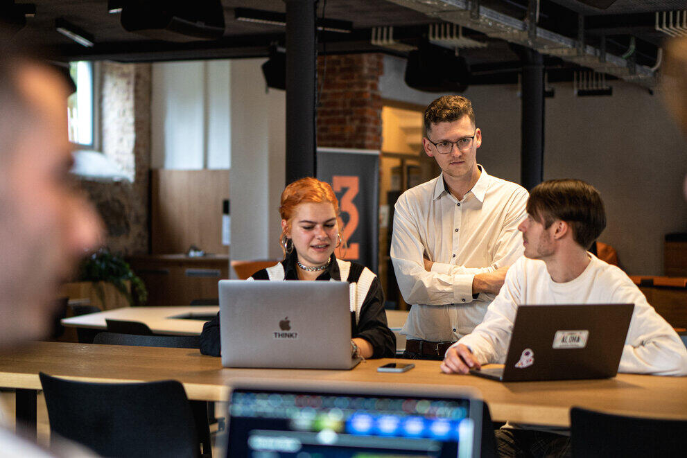 Three students are sitting in front of two laptops, chatting.