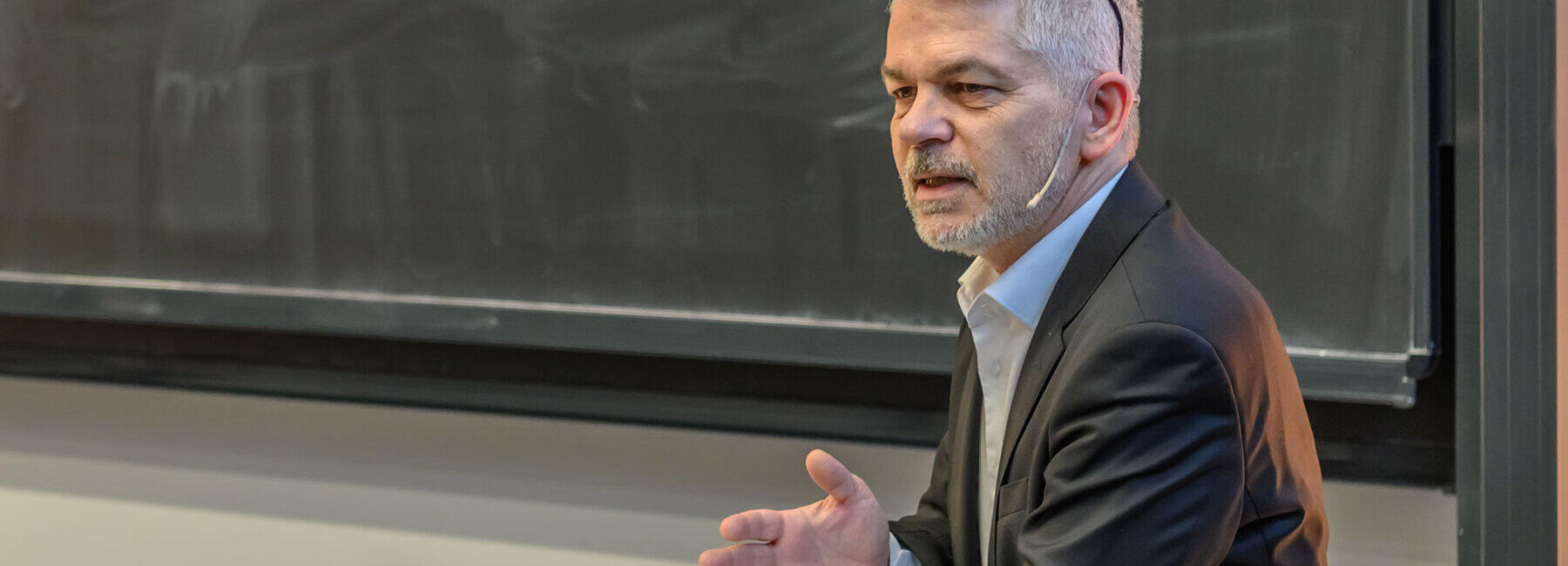A man with a determined expression stands in front of a poorly cleaned blackboard, his glasses pushed up into his hair, his left elbow resting on a lectern and his right hand open, speaking to an audience not visible in the picture.