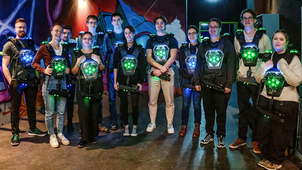 A group of 13 cheerful young people wearing laser tag vests and carrying phasers