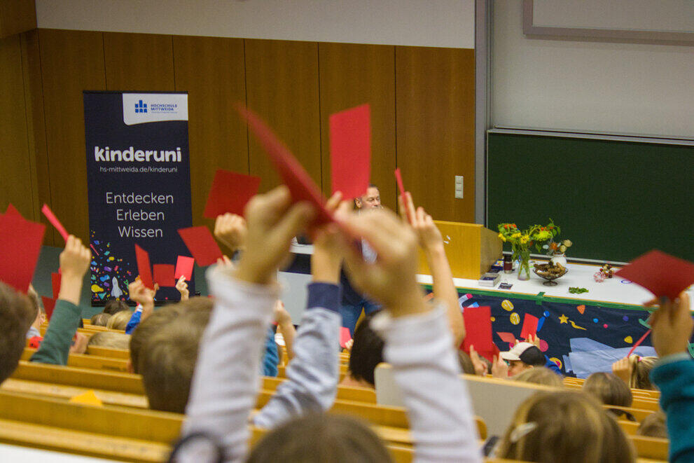 Kinder melden sich mit roten Karten im Kinderuni-Hörsaal. Vorn sieht man einen Aufsteller mit der Aufschrift „Kinderuni – Entdecken, Erleben, Wissen“