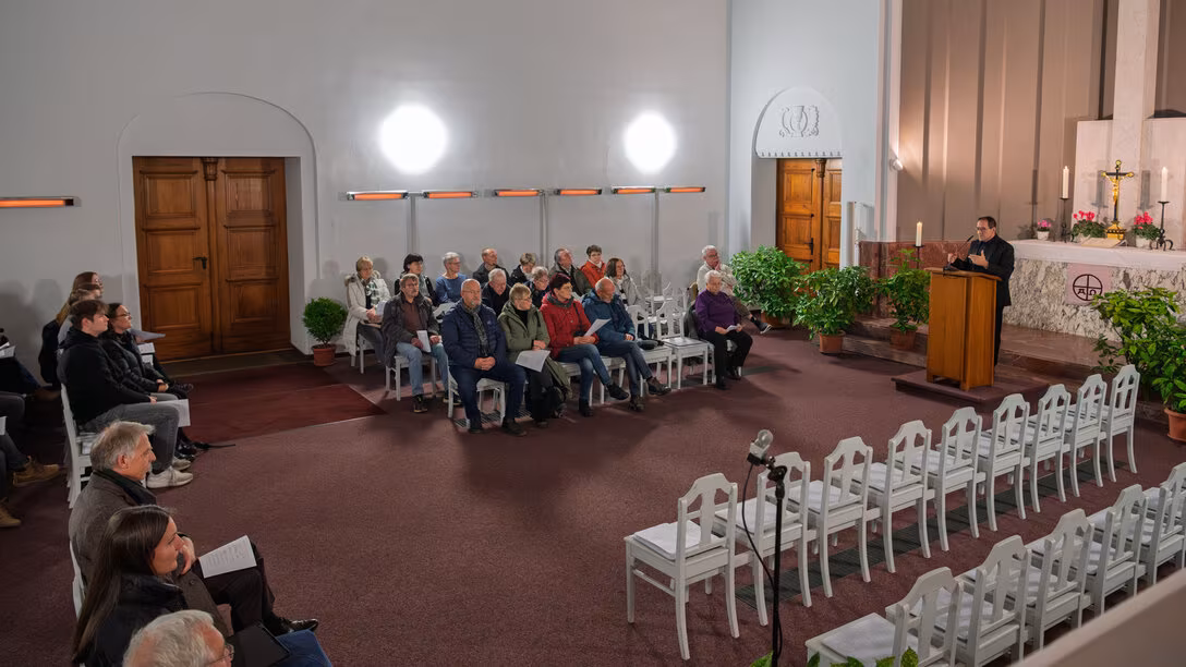 Blick in den großen Kapellenraum mit Besuchenden auf wei0en Stühlen. Am Rdnerpult vor inen Aktarr mit Kerzen und einem großen Kreuz eine männliche Person im Anzug.