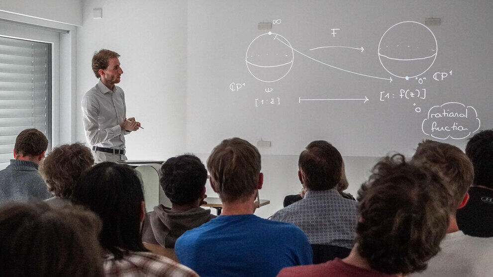 Männlicher Dozent vor projizierter Folie mit mathematischen Graphen. Im Vordergrund Zuhörende .