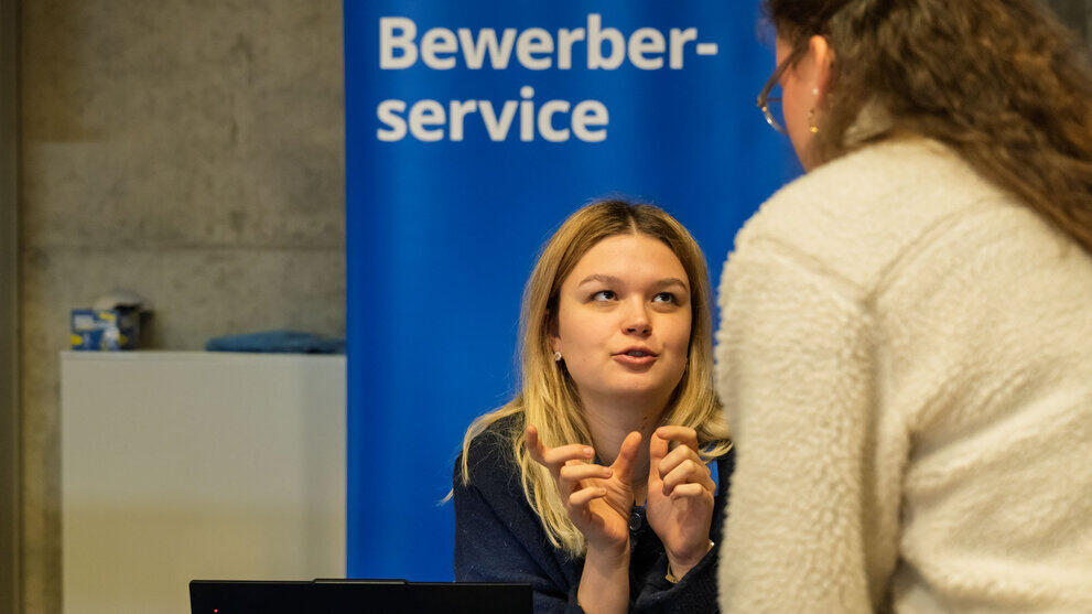 Junge Frau im Gespräch vor Rollup mit Aufschrift "Bewerberservice".