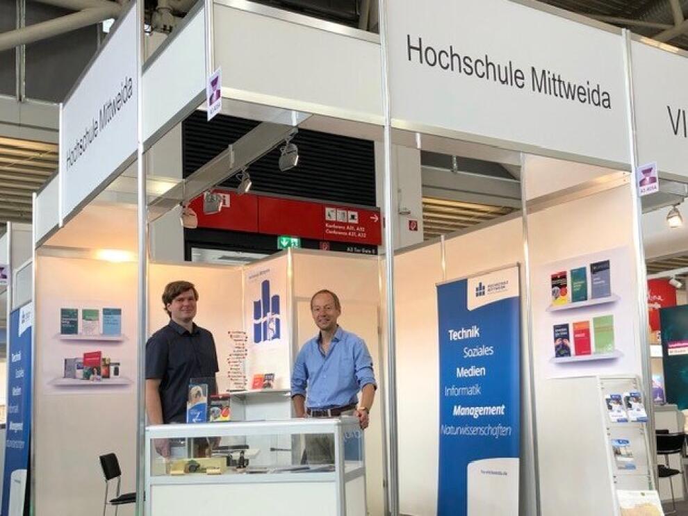 zwei männliche Personen stehen an einer Theke an einem Eck-Messestand mit Schriftbanner Hochschule Mittweida und Postern an den Wänden