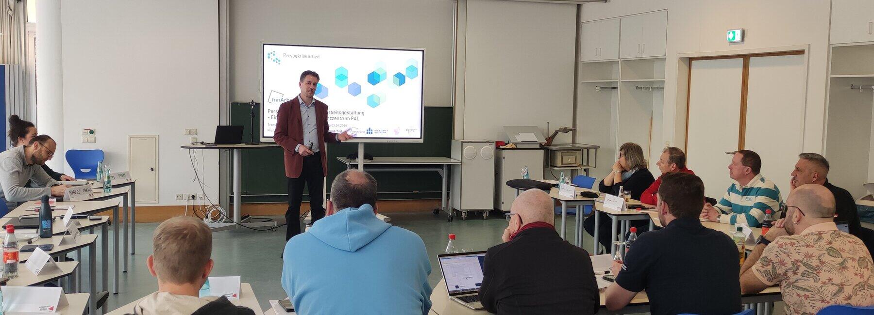 A man is standing in front of a presentation screen; other people are sitting at tables in a semicircle