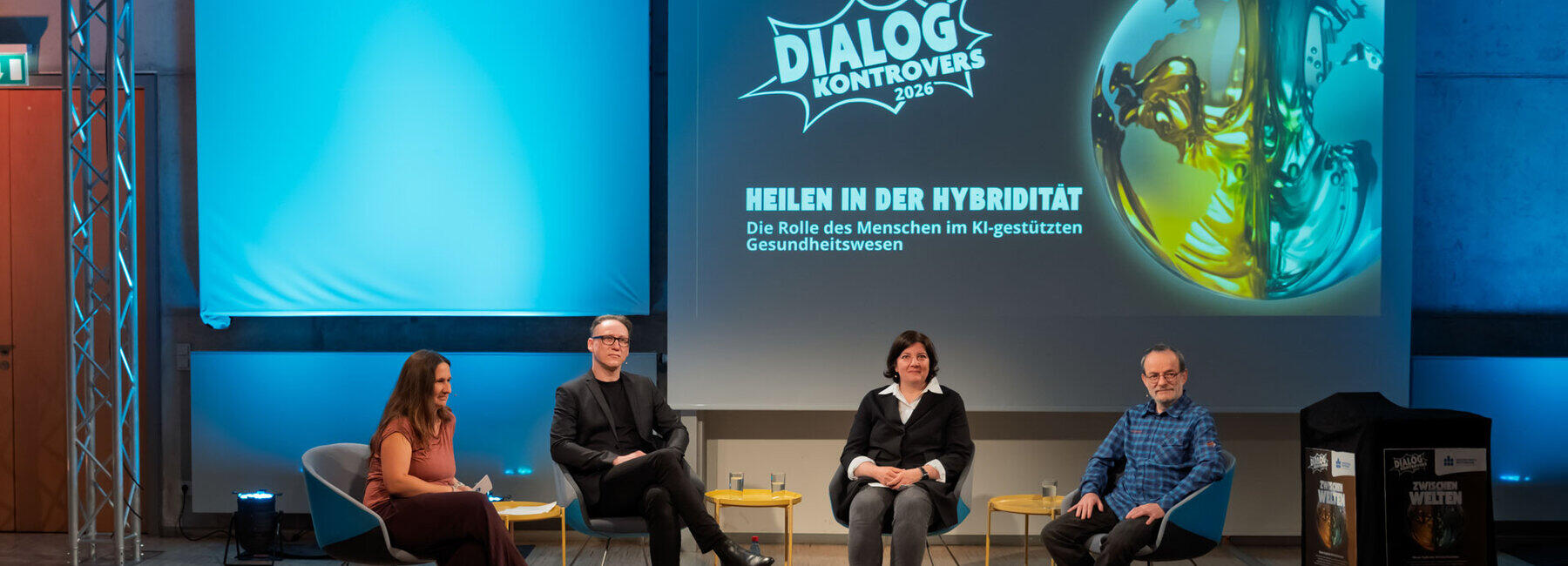 Zwei weibliche und zwei männliche Personen in Sesseln in einem studioähnlichen Raum. Auf der Leinwand hinter ihnen ist das Logo des Dialog Kontrovers zu sehen und das aktuelle Thema „Heilen in der Hybridität. Die Rolle des Menschen im Kl-gestützten Gesundheitswesen“ zu lesen.