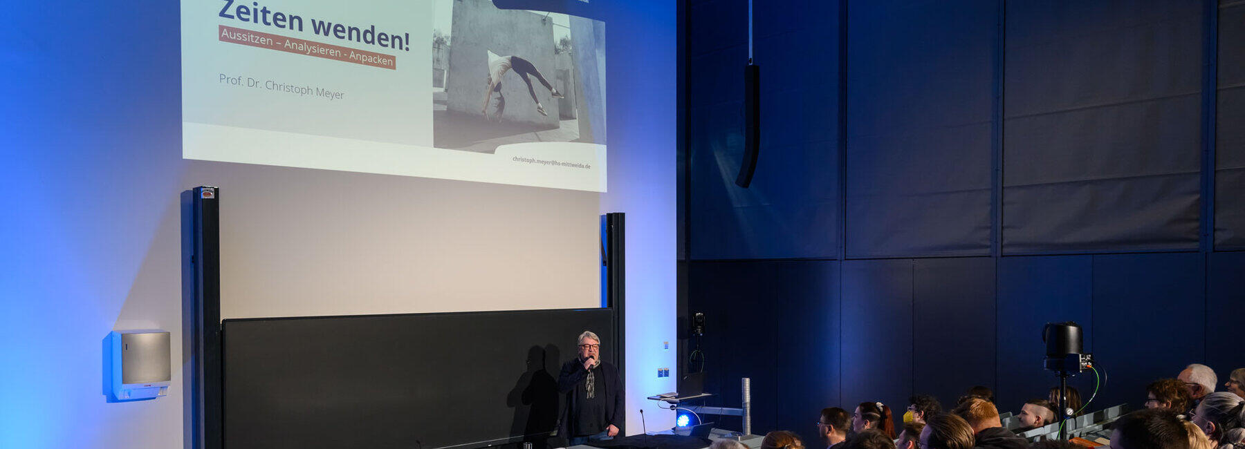 Seitlicher Blick von hinten in die ersten Reihen und auf Pult, Tafel und Beamerprojektion im Hörsaal. Projiziert ist die Begrüßungsfolie der Veranstaltung mit dem Text „Zeiten wenden! Aussitzen – Analysieren – Anpacken, daneben das Foto einer jungen Frau, die einen Rückwärtssalto vor einer grauen Betonwand macht.