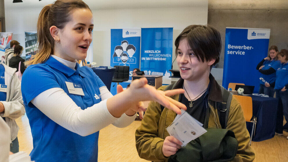 A young woman wearing a blue HSMW polo shirt, gesturing with her hands whilst talking to another young woman who is holding some leaflets. In the background, there is a roll-up banner bearing the words ‘Welcome to Mittweida’.