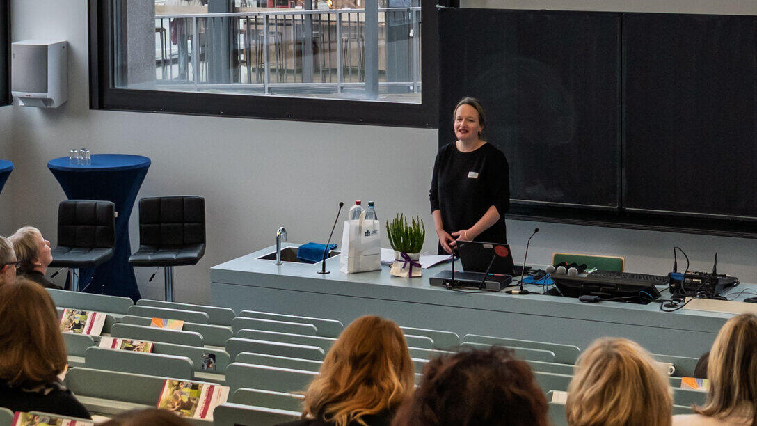 A woman smiling behind a lectern in a lecture theatre. In the foreground of the picture, the backs of people’s heads can be seen in the rising rows of lecture theatre seats.