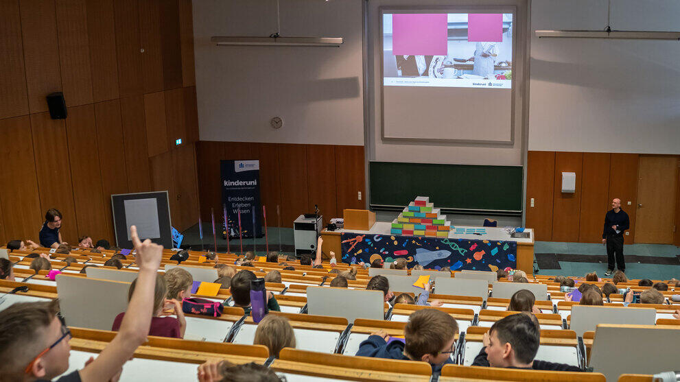 Blick von oben in den Hörsaal über die Köpfe der Kinder hinweg auf die Leinwand, die ein Foto mit zwei teilweise verdeckten Personen in einer Küche zeigt: eine sitzdend im Anzug, eine stehend mit Kochschürze.