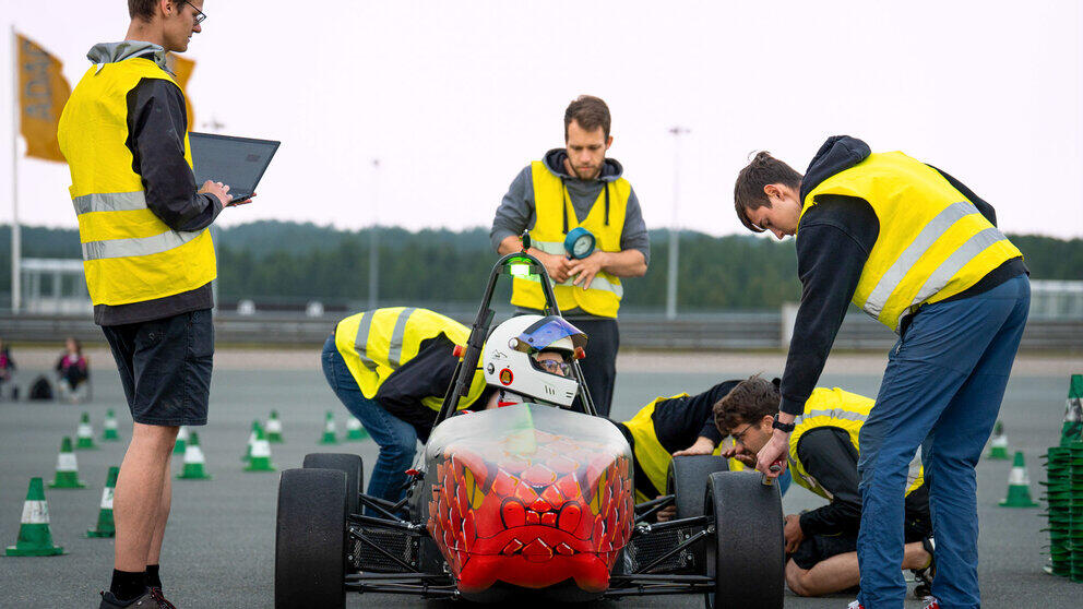 Five men wearing neon-yellow high-visibility vests are standing and kneeling around a red racing car, in which a person wearing a helmet is sitting. In the background, a section of track marked with red and white cones can be seen.