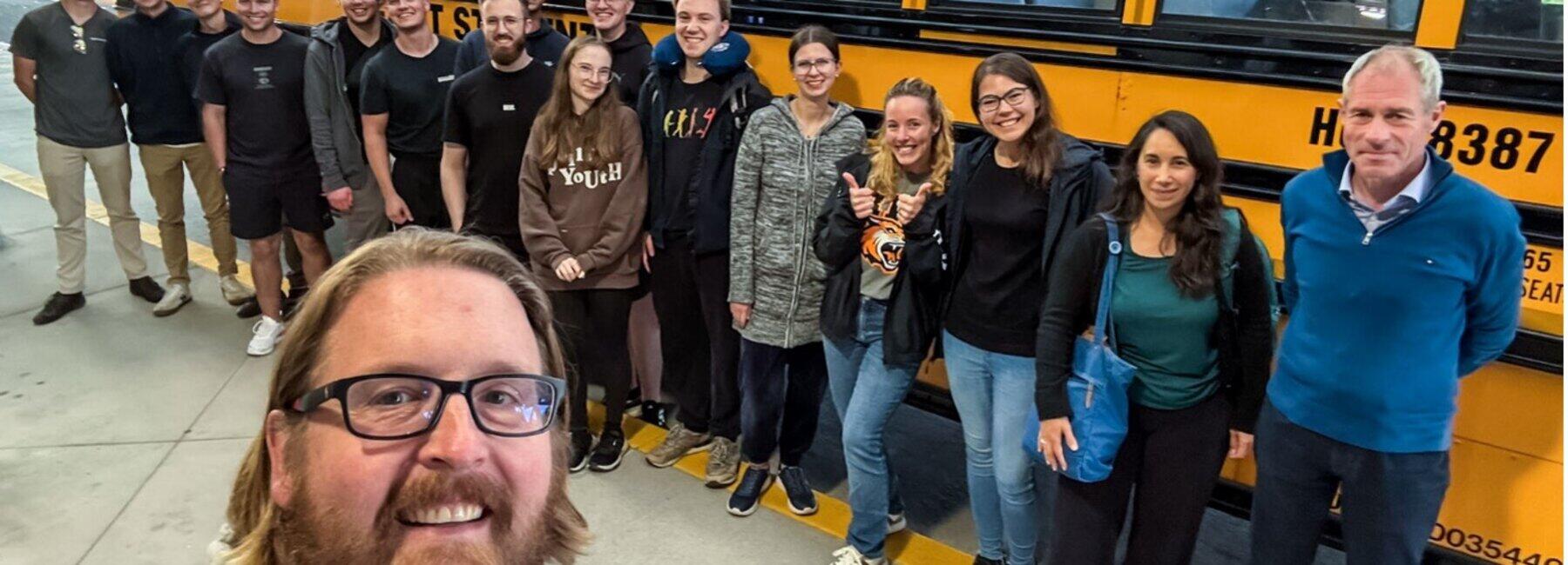 The selfie shows a group of mostly young people in front of a typical yellow American school bus. The photographer’s head can be seen in the foreground of the picture.