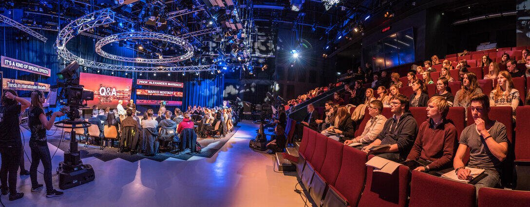 Panoramaansicht eines vollbesetzten Fernsehstudios mit Publikumsrängen.