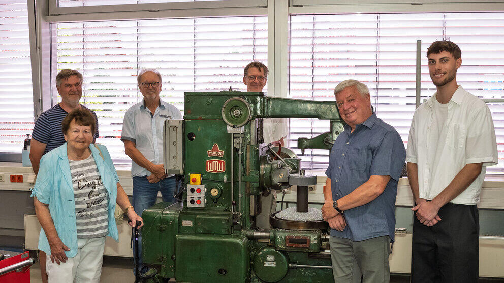 Six people are standing, smiling, around an old machine that is almost as tall as a man.