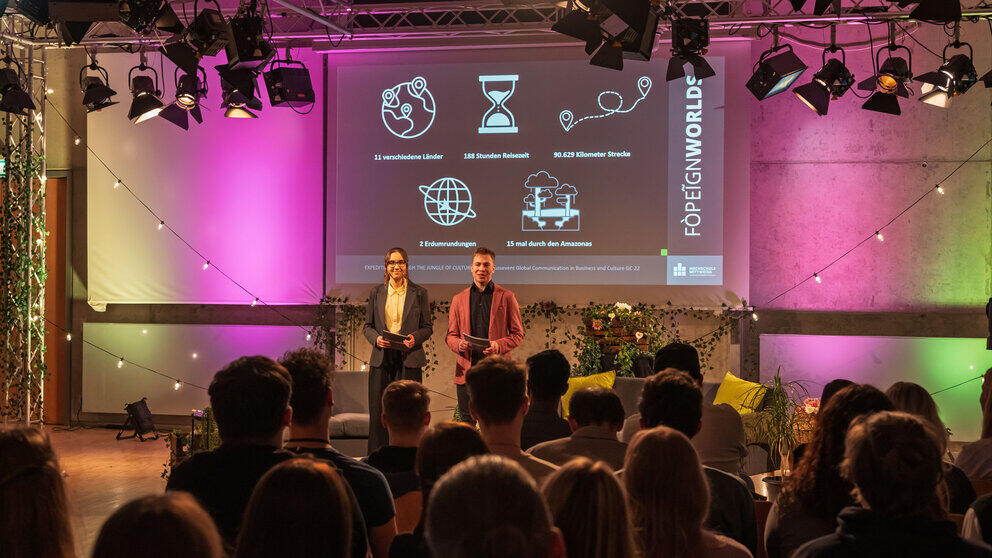 Two presenters – a young woman and a young man – are standing in the background of the studio set. In the foreground, the audience can be seen from behind. A slide containing graphics and corresponding figures is projected onto the screen behind the presenters: ‘11 countries, 188 hours’ travel time, 90,639 kilometres, 2 trips round the world, 15 times through the Amazon’.