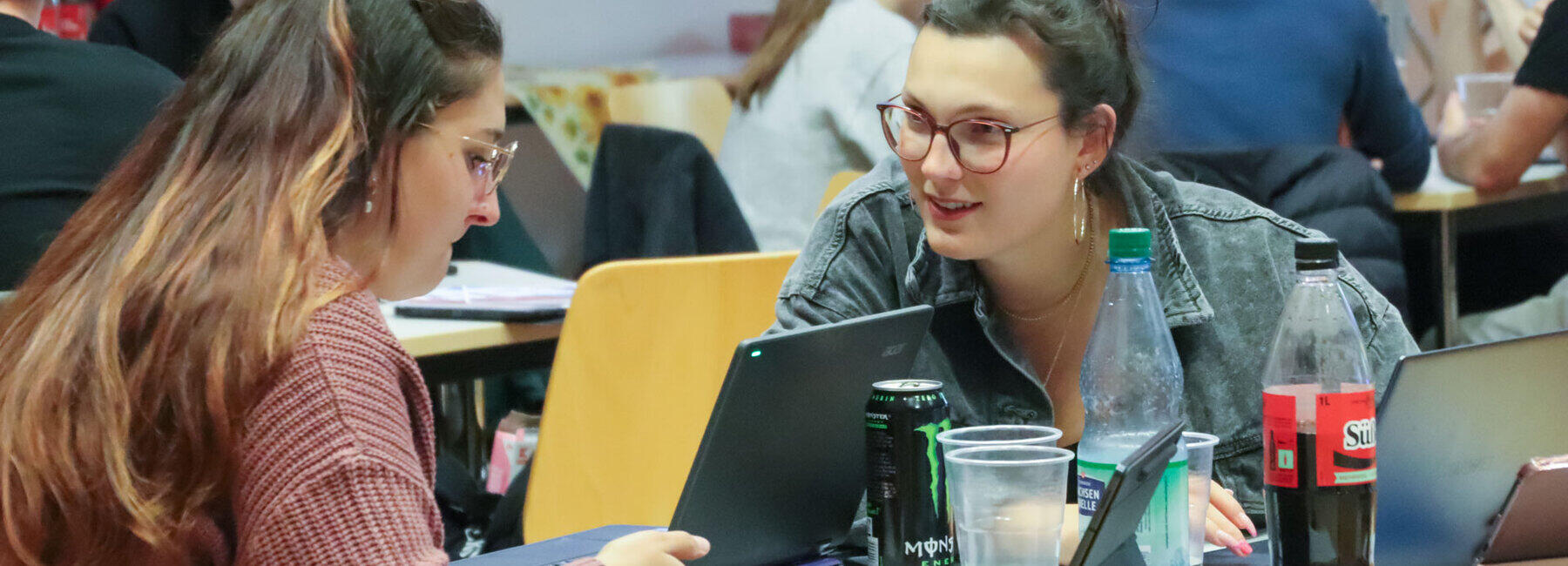 Two young women sitting at a table with laptops, mugs, bottles of cola and tins of energy drinks. The woman on the right is looking over at the one on the left with interest. In the background, other people can be seen sitting at tables.